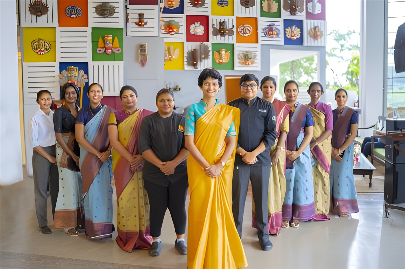 Women standing in a traingle formation in front of a colourful wall, many wearing yellow and pink and blue saris. These women run a hotel in Sri Lanka called Amba Yaalu Kandalama