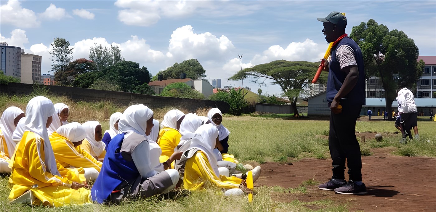 A group of girls sitting on a cricket pitch listening to a man talk.
