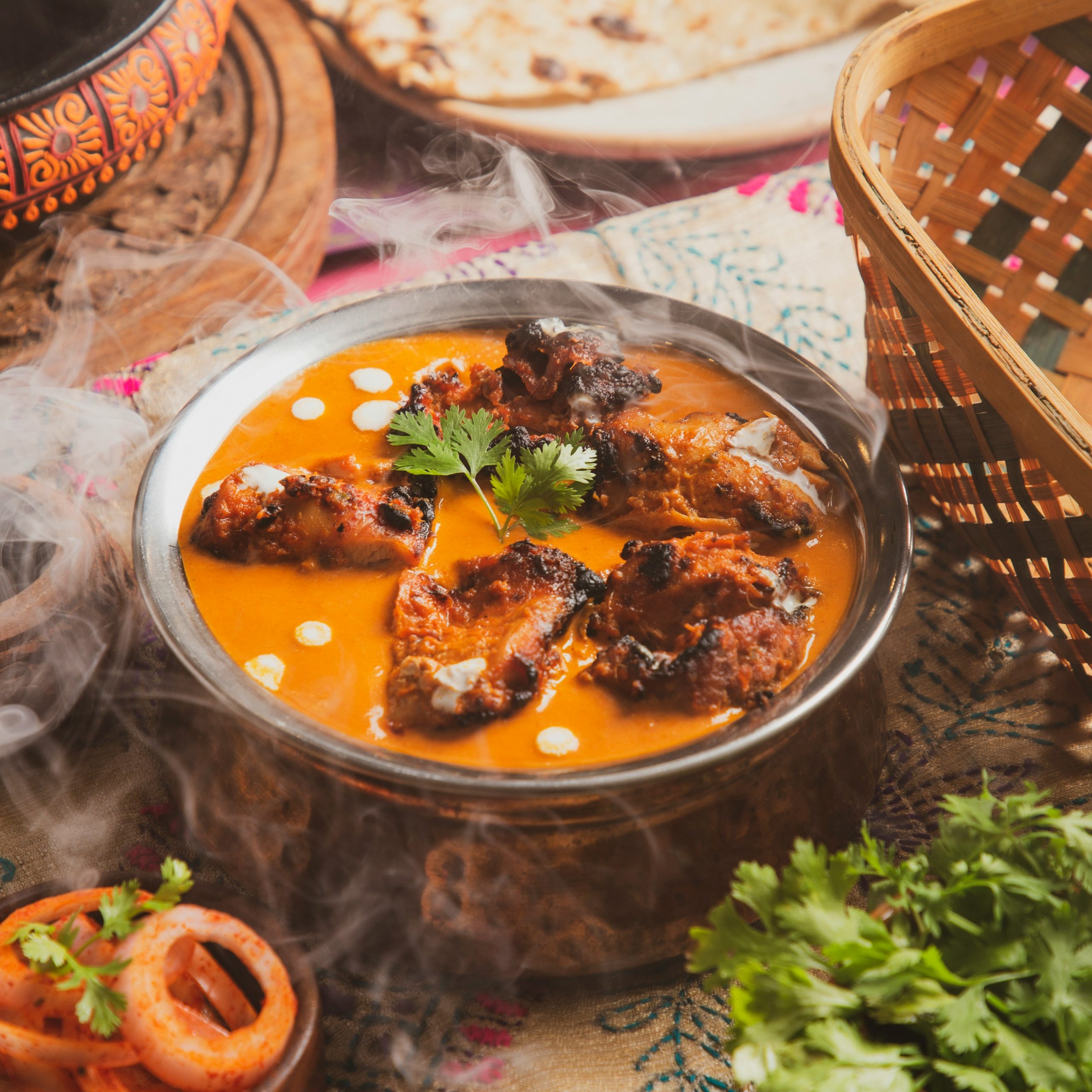 A steaming dish of butter chicken on a tabel set with baskets and other foods